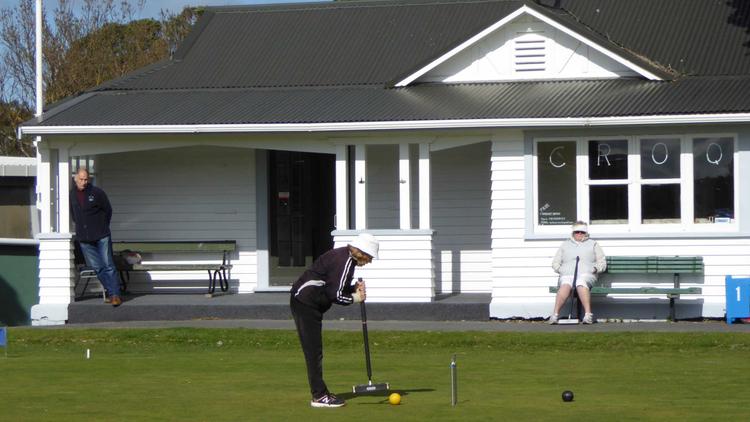 Photo of a person playing croquet outside of the Croquet Club, there is one person sitting on a bench watching and another person standing