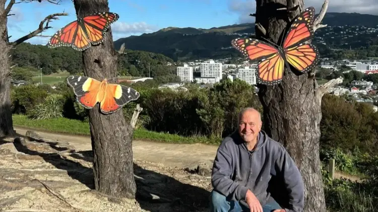 Artist Paul Forrest kneels in front of two trees with large butterfly sculptures attached to the trunks.