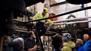 Crowd gathers inside a ship, watching a man talking from a ladder