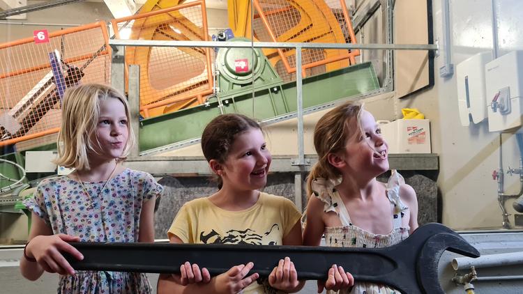Three children holding a large wrench between them, cable car winding gear in the background