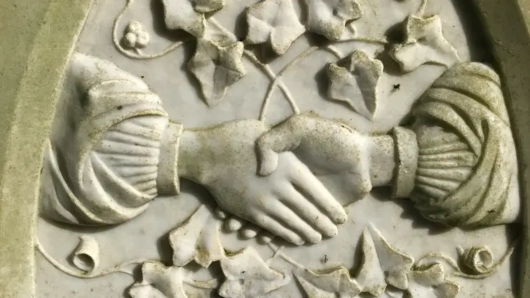 An aged white marble headstone carved with the image of two hands clasped together surrounded by ivy vine motifs.