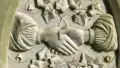 An aged white marble headstone carved with the image of two hands clasped together surrounded by ivy vine motifs. 
