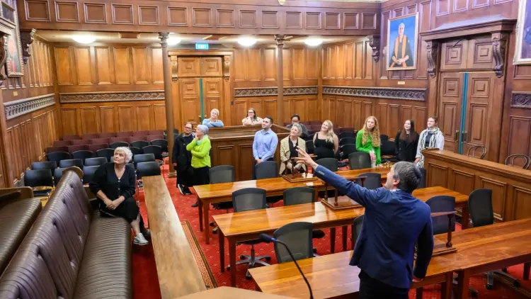 a group of people are in a historic courtroom, a man in a suit stands at the front gesturing for the guests to look at a portrait 