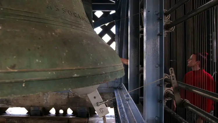 Photo of one of the large hanging bells, larger than a person in the Carillon Tower. A visitor admires it through a protective barrier.