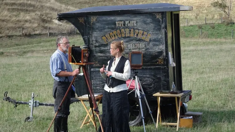 Two people stand next to a small trailer reading "Wet Plate Photographs" in vintage font.