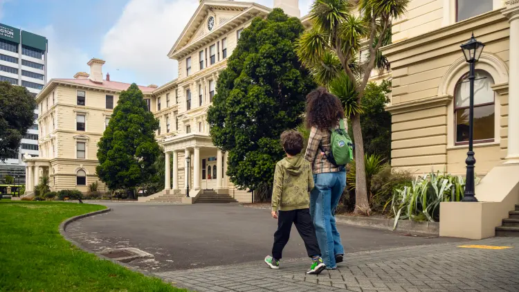 Two children walking in front of a yellow stone building, green grass on the left and trees in the background