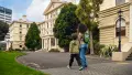 Two children walking in front of a yellow stone building, green grass on the left and trees in the background