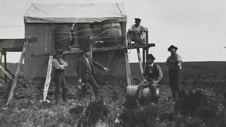 Black and white photograph of men standing in front of a shed and barrels