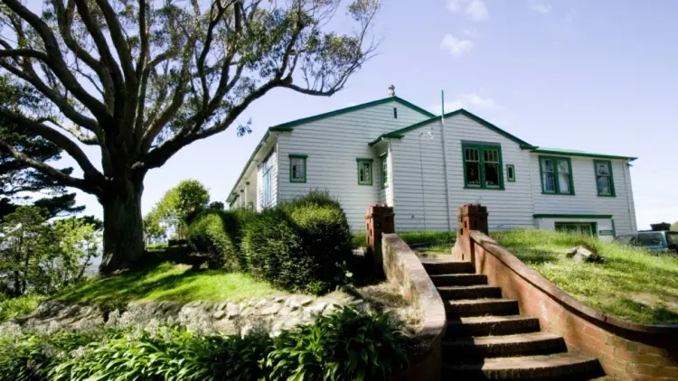 Colour photograph of a white house in the background with brick stairs leading up, and a tree on the left.