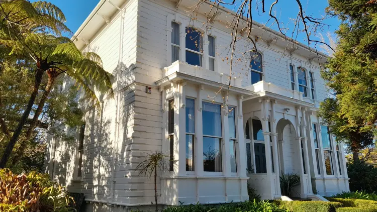 Colour photograph of a cream coloured wooden house surrounded by green foilage.