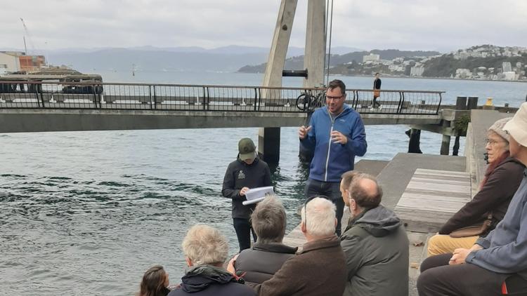 Photo of multiple people of various ages sitting on the side of the waterfront listening to a person presenting. 