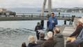 Photo of multiple people of various ages sitting on the side of the waterfront listening to a person presenting. 