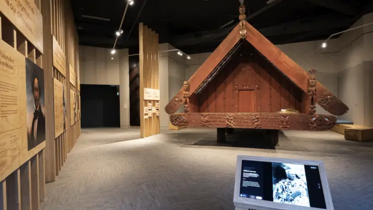 Inside a museum, a beautifully carved Māori pātaka stands surrounded by interpretation signs.