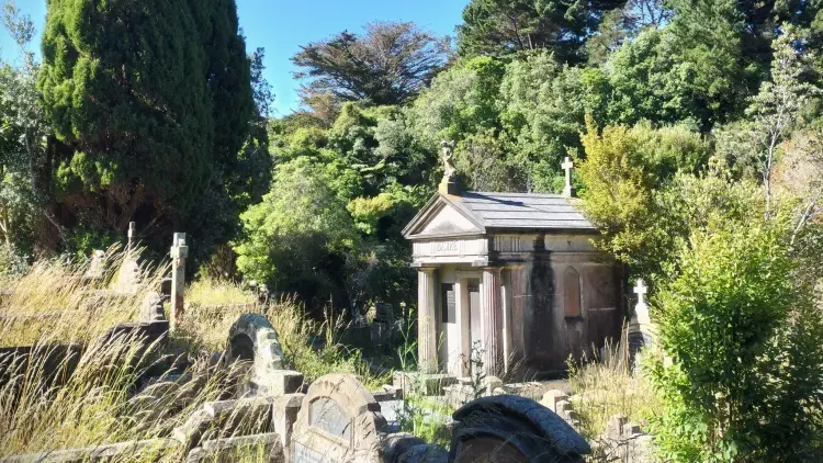 A photo of a cemetery in daytime. Large overgrown bushes surround gravestones and a large white mausoleum stands in the center frame.