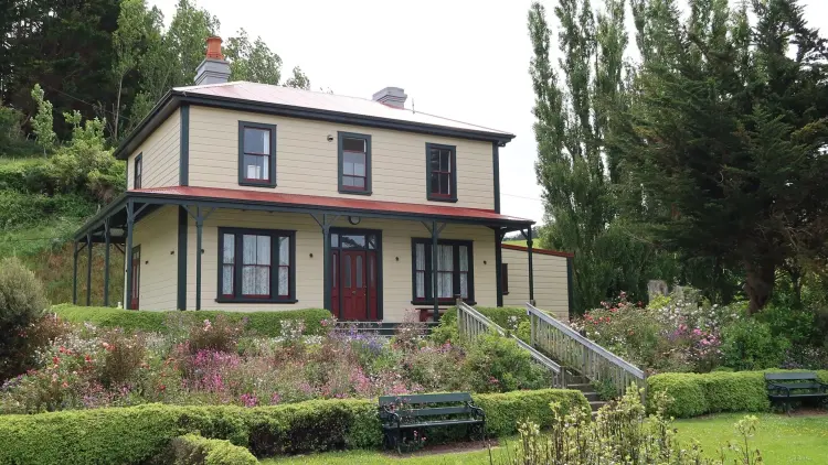 Photograph of a cream coloured 2 story house surrounded by a small garden containing hedges, pink flowers and trees.
