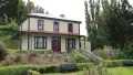 Photograph of a cream coloured 2 story house surrounded by a small garden containing hedges, pink flowers and trees.