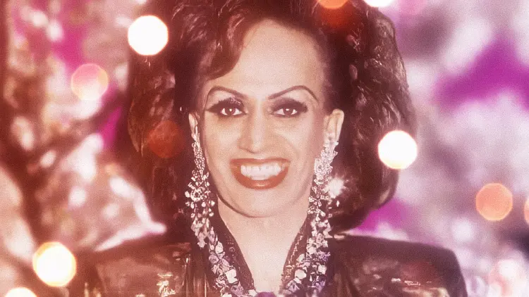 A photograph of Carmen Rupe, smiling wide and illuminated by glittering light.