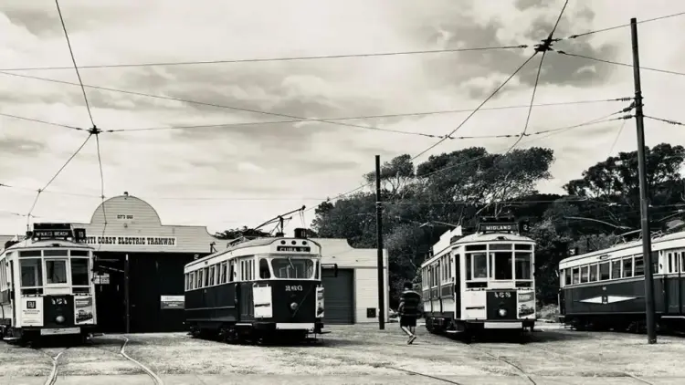 Historic photo of 4 trams, with power lines overhead, outside a shed