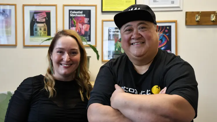 Two people smiling at the camera with framed posters on wall behind