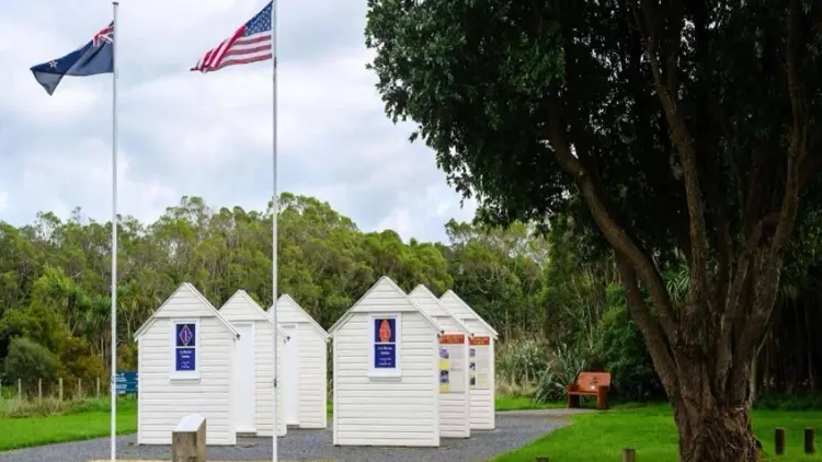 Outdoor photo of two flag poles and mock recreations of military huts. The New Zealand and American flags are flying.