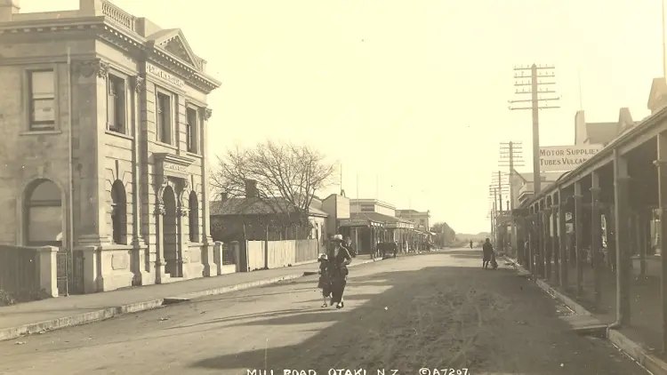 Sepia toned historic photo of a woman and child walking down Ōtaki Main Street 1923. Text at bottom reads "Mill Road Otaki NZ ©A7207"