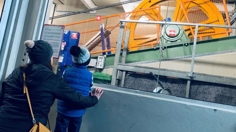 A child and adult in beanies look through a window to a large yellow winding wheel and machinery.
