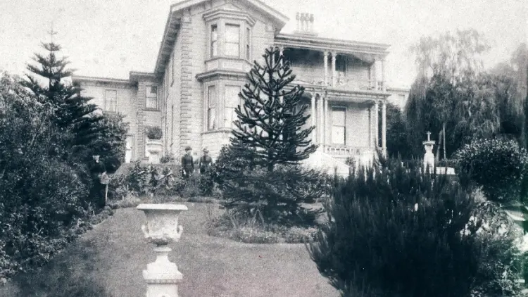 Black and white image of a two story colonial house and a garden with pine trees