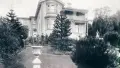 Black and white image of a two story colonial house and a garden with pine trees