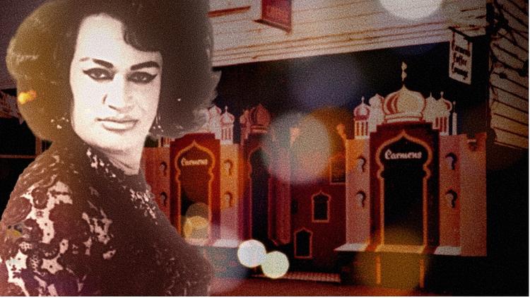 Image of Carmen Rupe staring into the camera with a gallery in the background. 