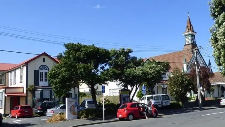 Colour photograph of buildings and a tree, red car parked on the street