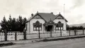Sepia photograph of a house with dark roof and white picket fence, flag pole